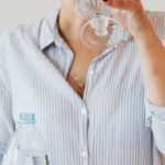 Smart Water Bottle - Female with short hair in eyewear and shirt with stripes standing near white table and drinking while holding bottle of water