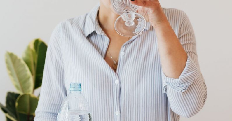 Smart Water Bottle - Female with short hair in eyewear and shirt with stripes standing near white table and drinking while holding bottle of water